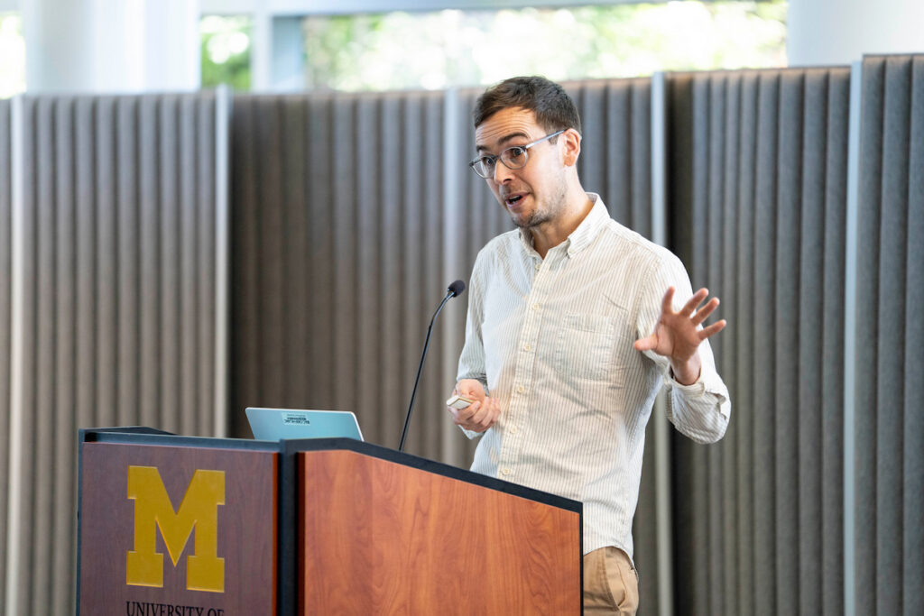 A person with glasses stands at a podium featuring the University of Michigan logo, gesturing while speaking into a microphone.