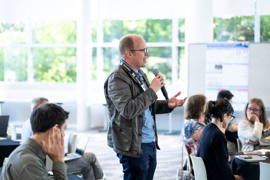 A person stands speaking into a microphone at a conference, addressing an audience seated at tables.