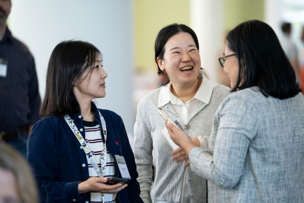 Three individuals engage in a cheerful conversation at an event, with one person laughing heartily while holding a phone.