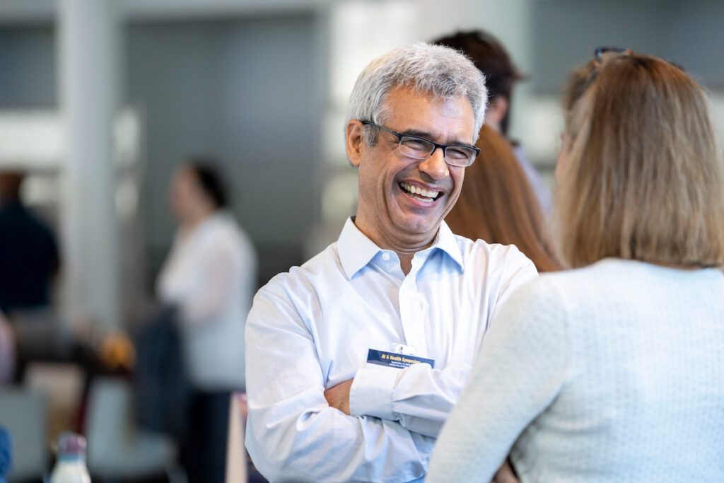 A person with gray hair and glasses laughs heartily while engaged in conversation with another individual at an event.