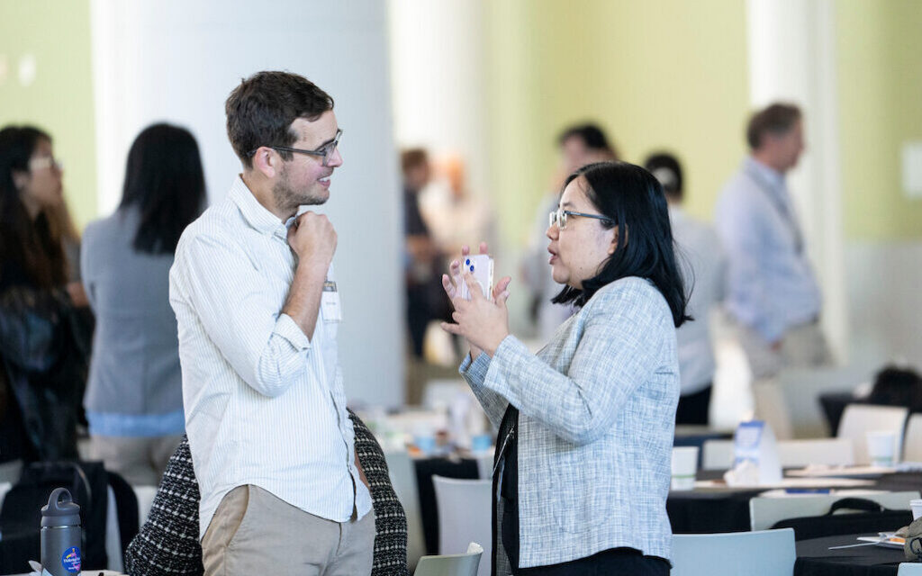 Two individuals engage in conversation at a conference, with one holding a smartphone while the other adjusts their collar.