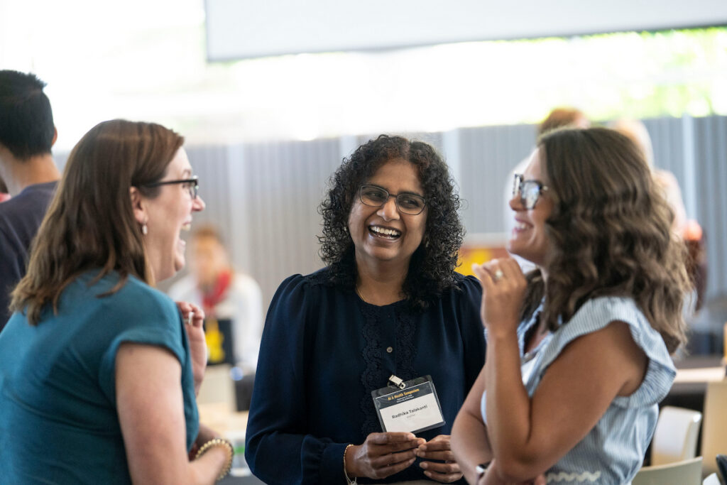 Three individuals engage in a lively conversation at an event, with one smiling broadly while holding a name badge.