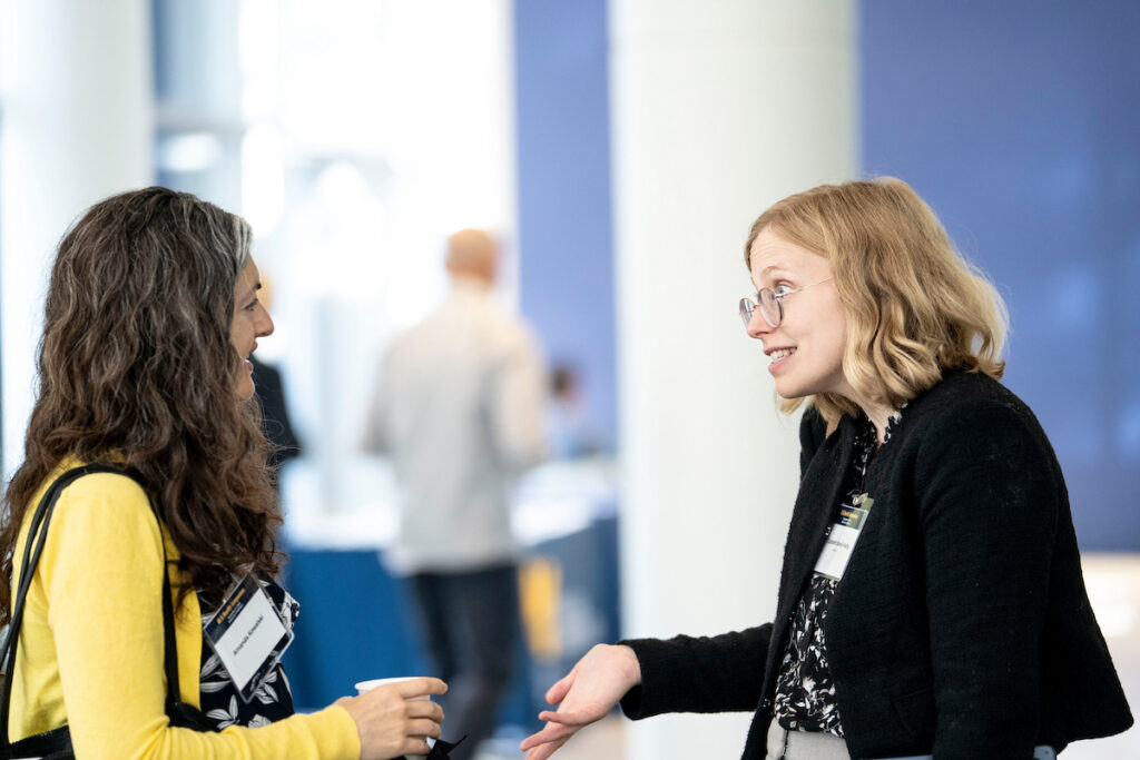 Two individuals engage in conversation at a conference, with one gesturing while holding a cup and the other smiling attentively.