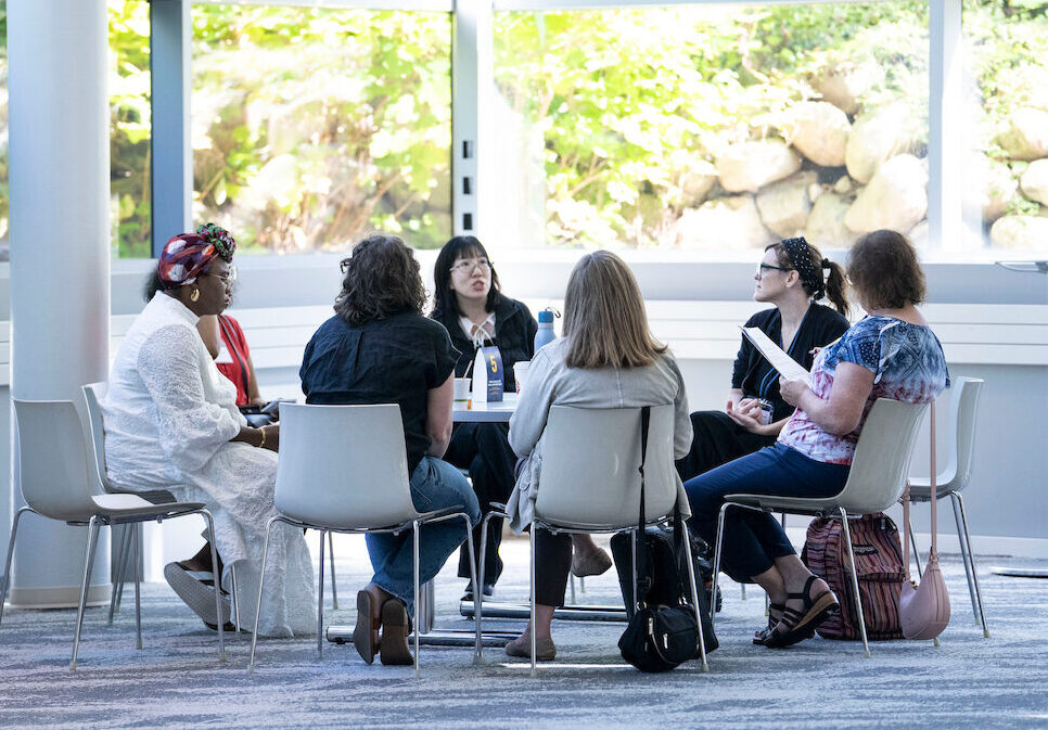 A group of individuals sits in a circle around a table in a bright, modern room with large windows overlooking greenery, engaged in conversation.
