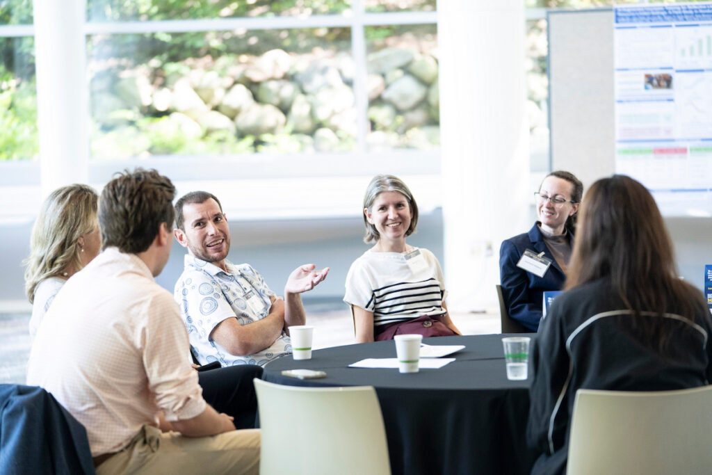 A group of people engage in lively conversation around a table in a bright, modern room with large windows.