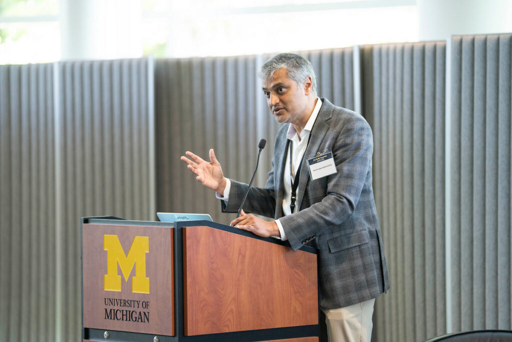 A person in a suit stands at a University of Michigan podium, gesturing while speaking into a microphone.
