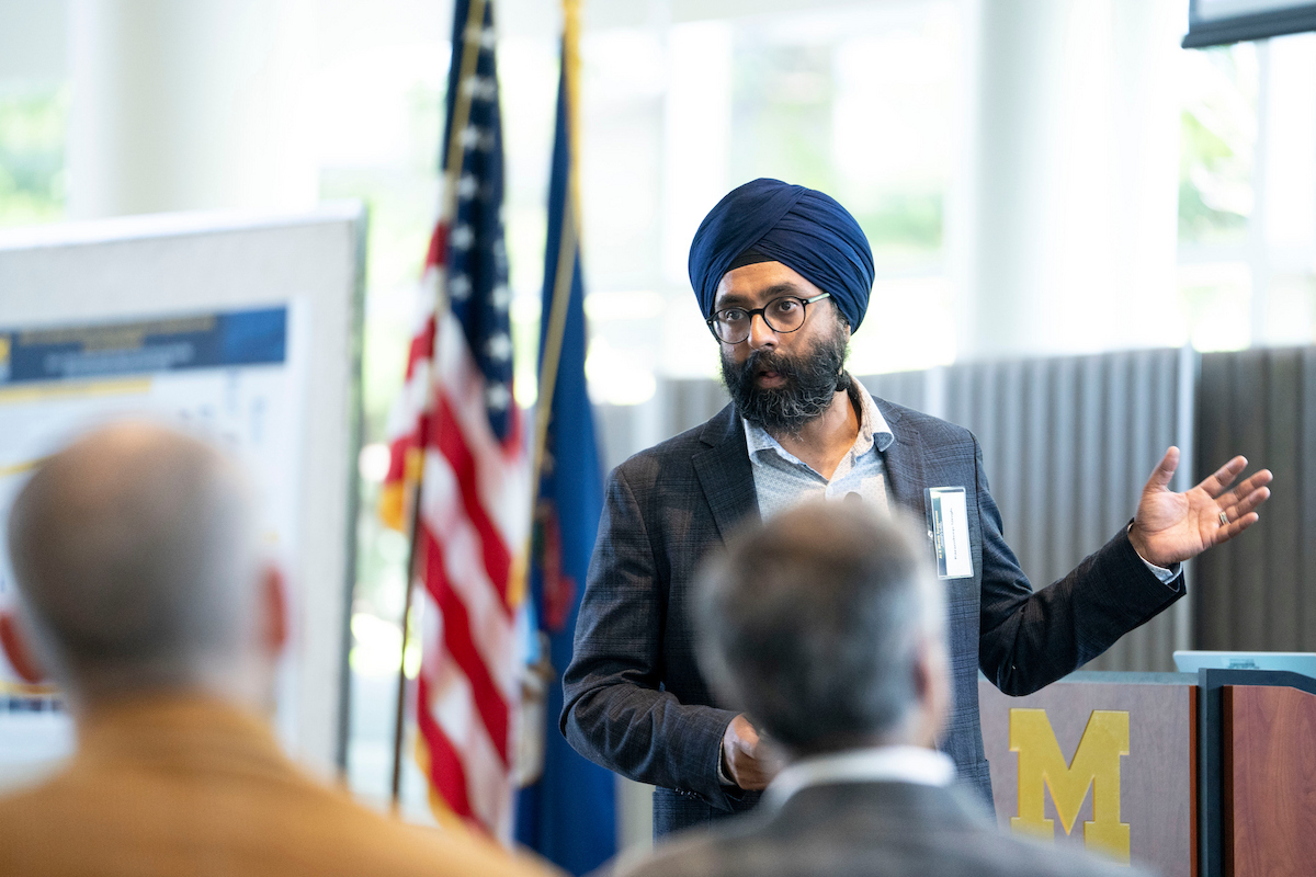 A person wearing a turban and glasses stands at a podium, gesturing while speaking to an audience in a room with flags and a large "M" logo.