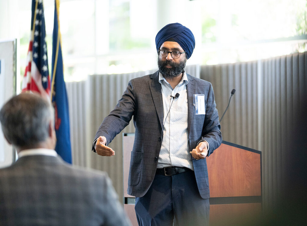 A person wearing a turban and glasses stands at a podium, gesturing while speaking to an audience in a well-lit room with flags in the background.