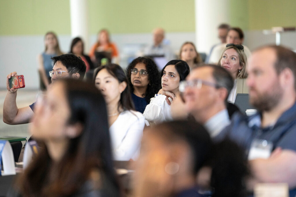 A diverse group of attendees at an event, some taking photos and others listening attentively.