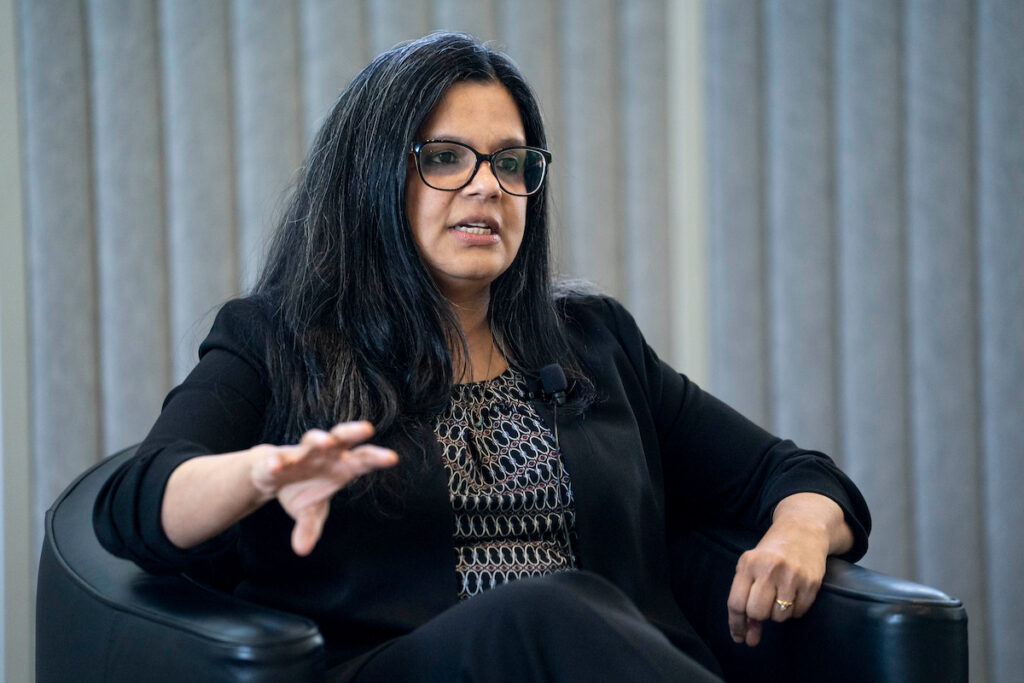 A person with long dark hair and glasses sits in a black chair, gesturing while speaking, against a backdrop of vertical blinds.