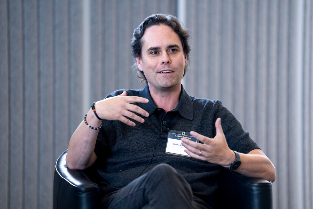 A person sits in a black leather chair, gesturing while speaking, wearing a dark polo shirt and a name badge.