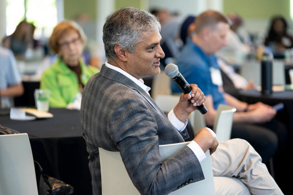 A person in a suit speaks into a microphone while seated at a conference table, with other attendees visible in the background.