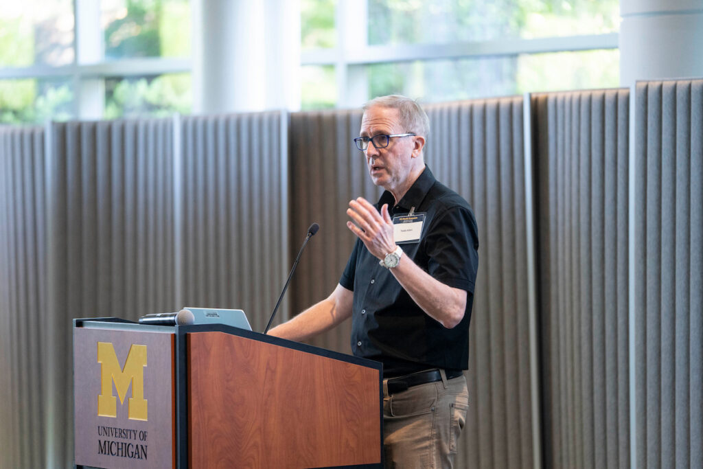 A person in a black shirt stands at a University of Michigan podium, speaking into a microphone while gesturing with their hands.