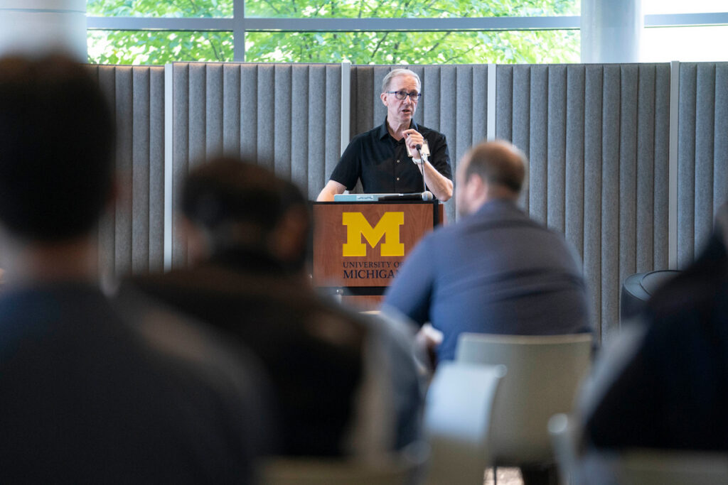 A person speaks at a podium bearing the University of Michigan logo, addressing an audience in a modern conference room.