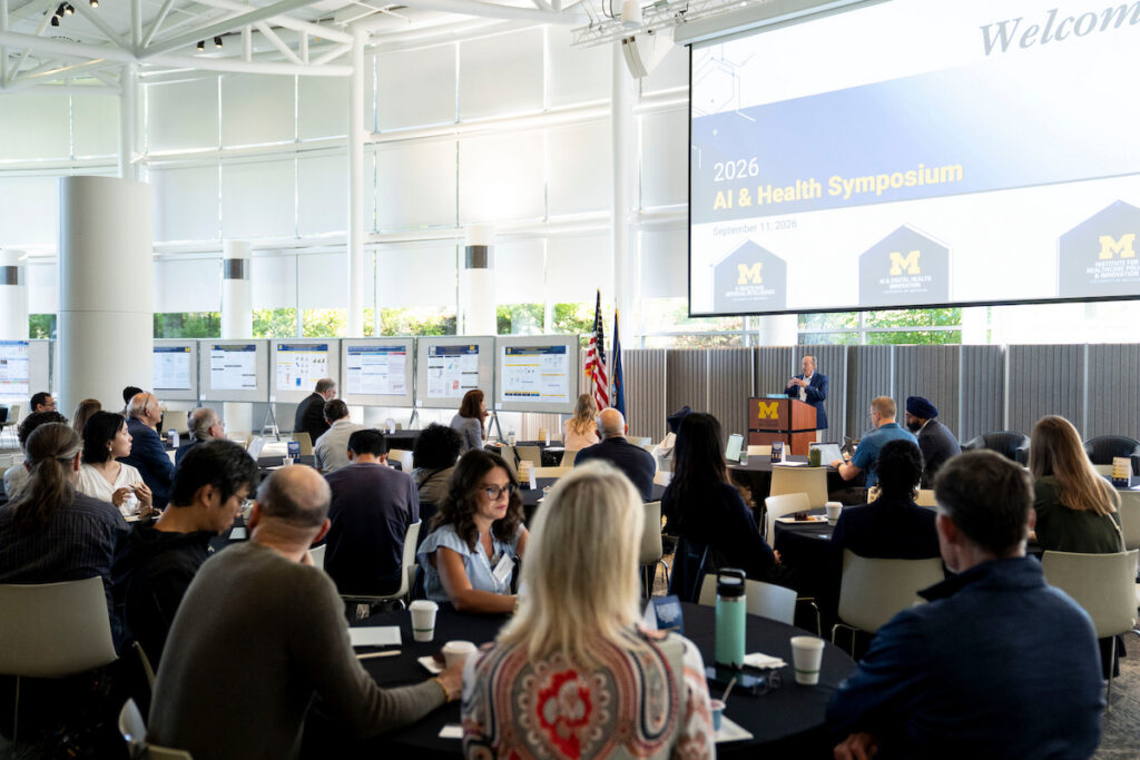 Attendees gather at round tables in a modern conference hall during the 2026 AI & Health Symposium, listening to a speaker at a podium beneath a large screen displaying the event title and date.
