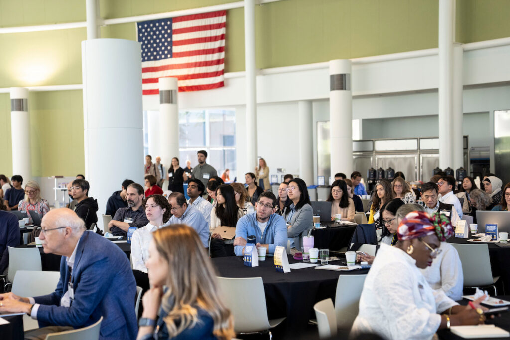 A diverse group of attendees sits at tables in a large room, attentively listening to a presentation under an American flag.