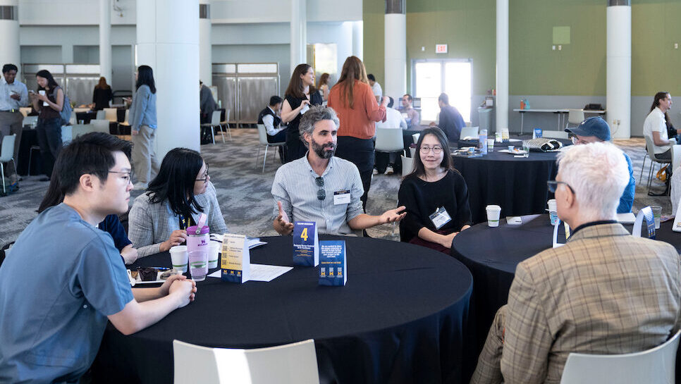 A group of individuals engage in conversation around round tables in a spacious, modern conference hall.