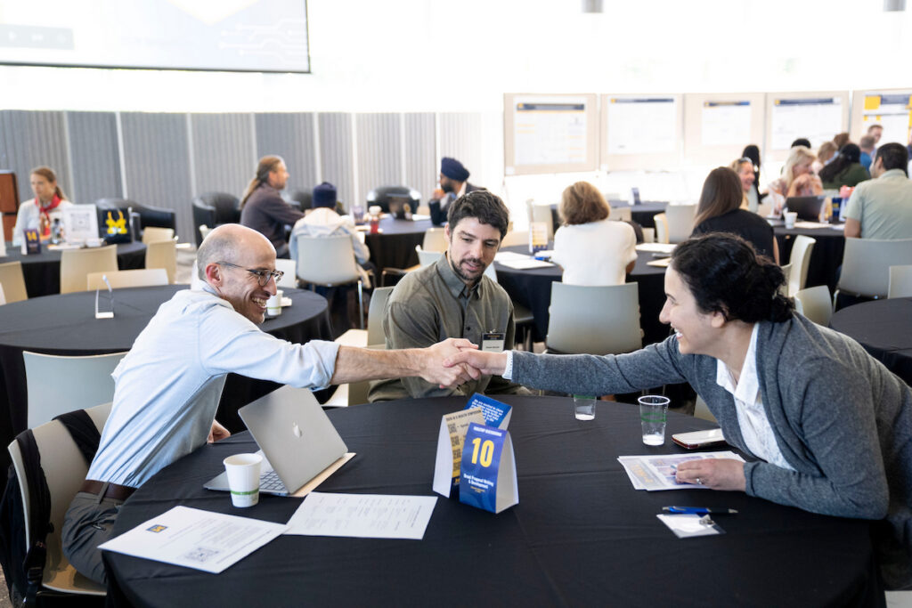 Two individuals shake hands at a conference table while others engage in conversations in the background.