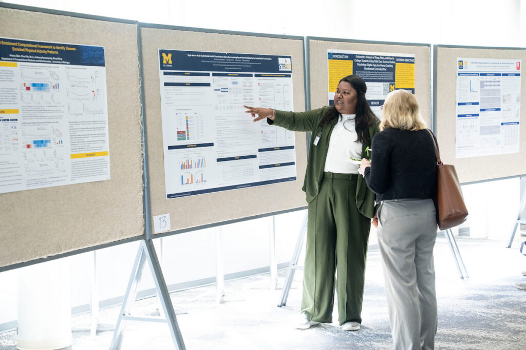 Two individuals engage in conversation at a research poster session, with one person pointing to details on a scientific display.