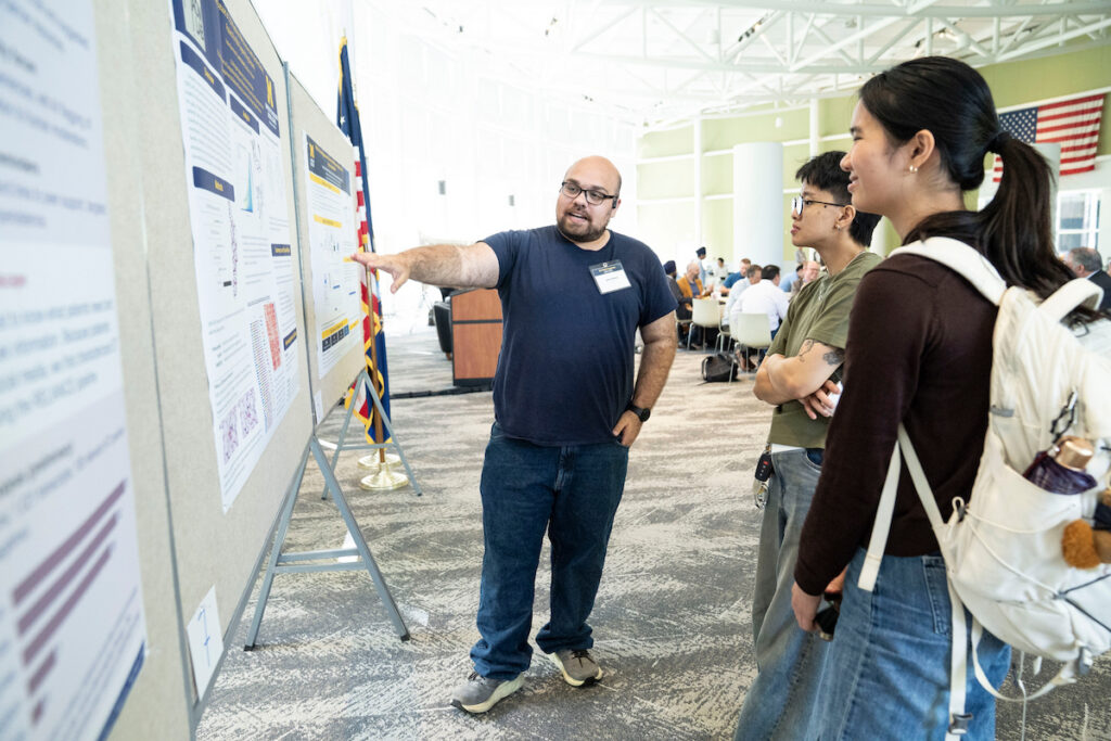 A person gestures toward a research poster while two others observe attentively at an academic conference.