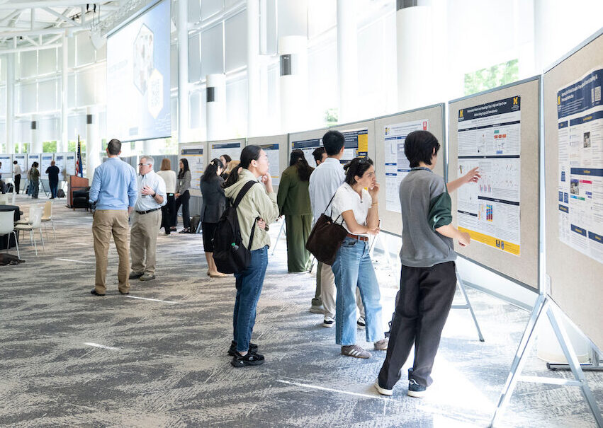 Attendees at a conference engage with research posters in a bright, modern hall with high ceilings and large windows.