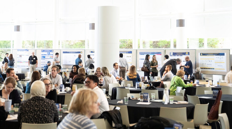 A diverse group of attendees engages in conversation and networking at a conference or exhibition held in a bright, modern hall with large windows and display boards.