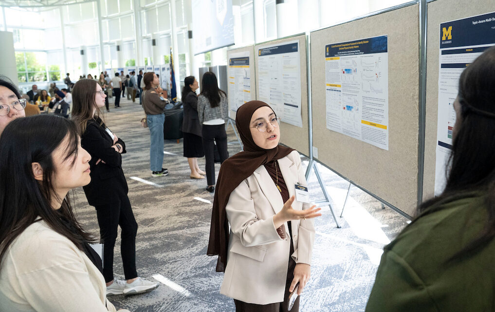 A person in a beige blazer and brown headscarf gestures while explaining a research poster to an audience at an academic conference.