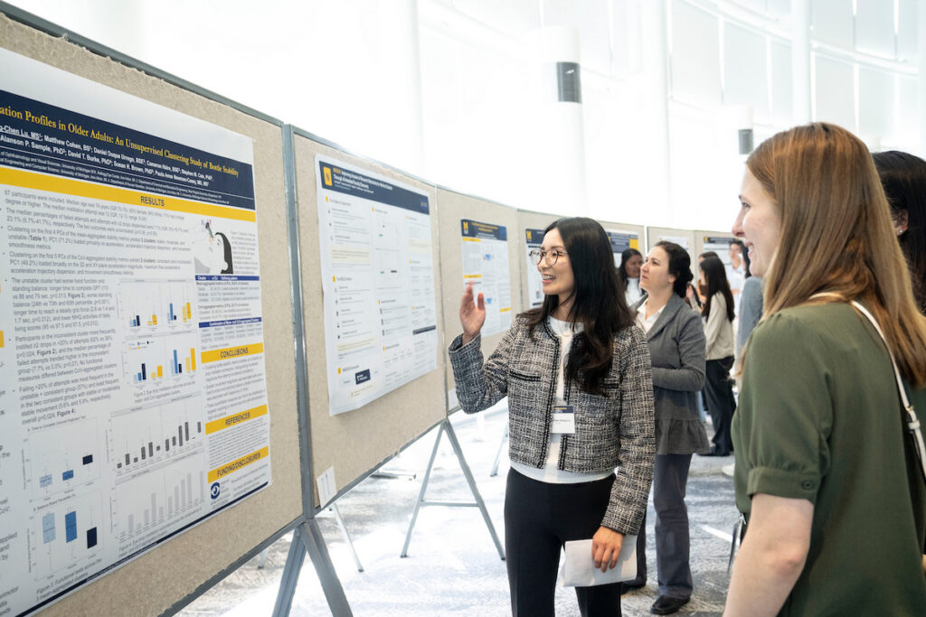 A person presents a research poster at an academic conference while engaging with an attendee.