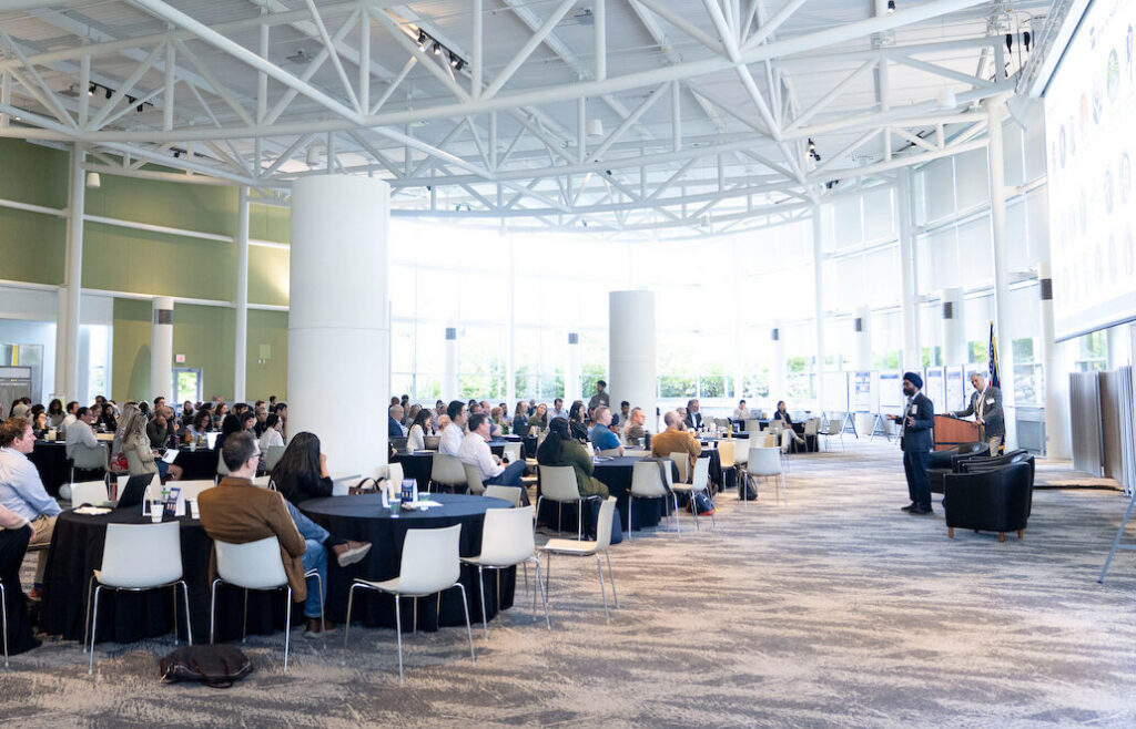 A large gathering of people seated at round tables in a modern, spacious hall with a high, exposed-truss ceiling and large windows.