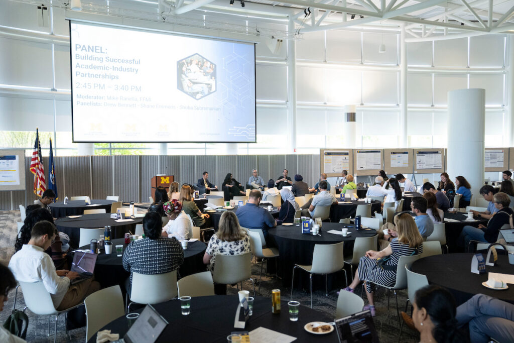 A panel discussion on building successful academic-industry partnerships is taking place in a modern conference room with attendees seated at round tables.